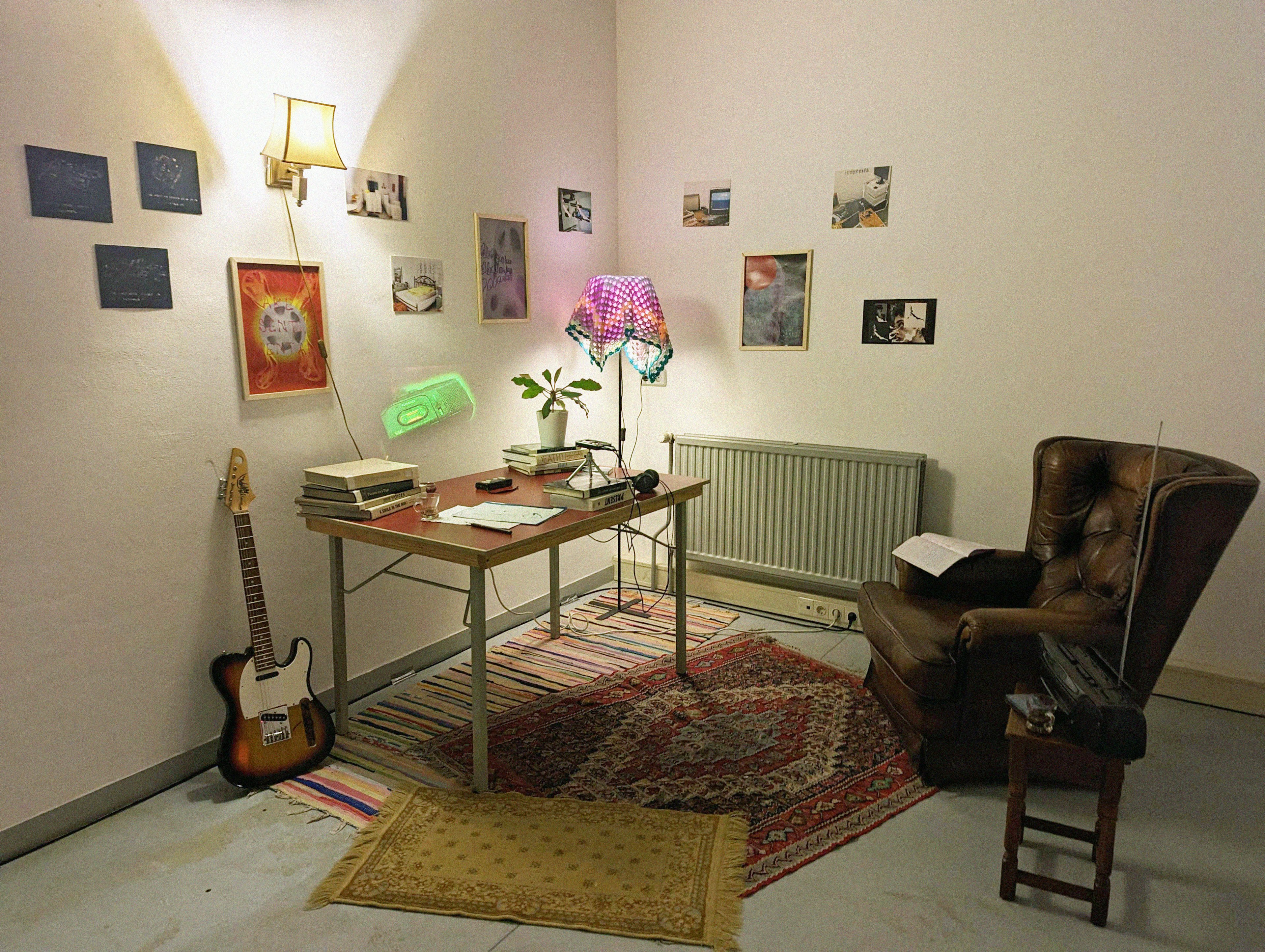 A domestic setting featuring rugs, an arm chair near a radio, a desk with books and a tape recorder, a guitar, and a wall decorated with several photos, a sconce, and a small projection of a video