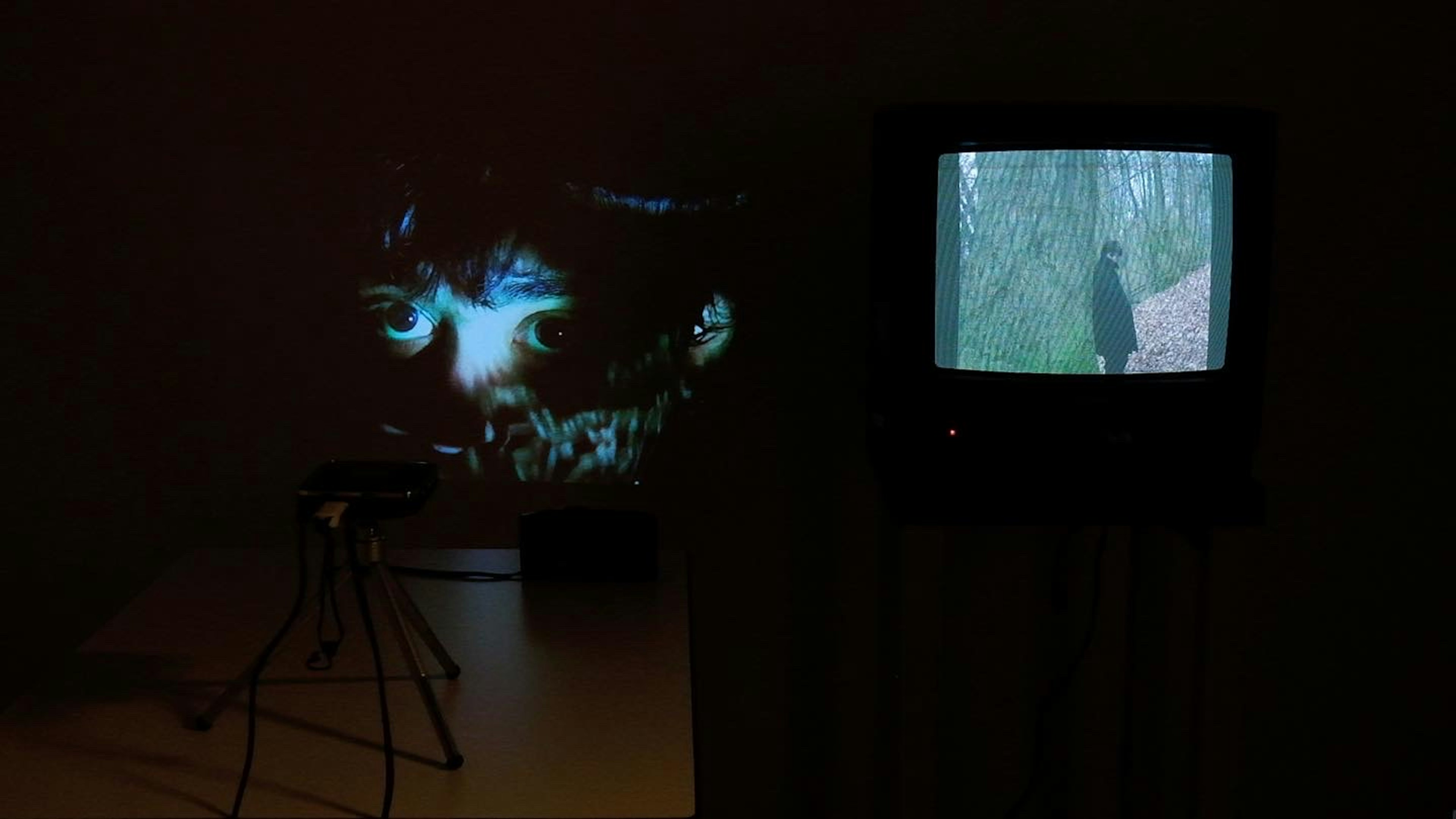A dark wall with a projection on the left, and a CRT television on the right. The projected video features Milo's face under a light image of a ghost. The video on the CRT television features a cloaked figure in a forest setting. 